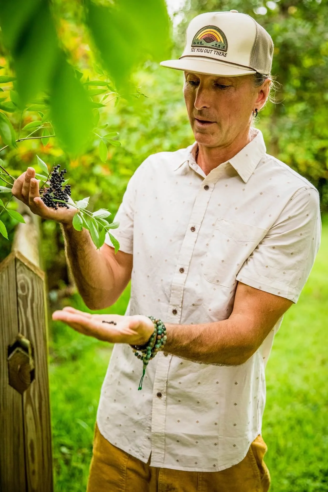 A man wearing a white button-up shirt, yellow pants, and a beige cap with rainbow and sunset embroidery, holding a branch of ripe elderberries in an outdoor setting surrounded by green foliage.