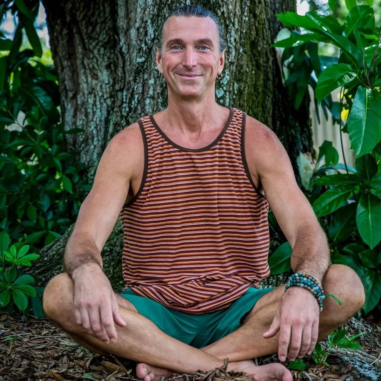 A man with long hair, wearing a striped tank top and shorts, sitting cross-legged outdoors near a large tree, smiling, with green foliage in the background.