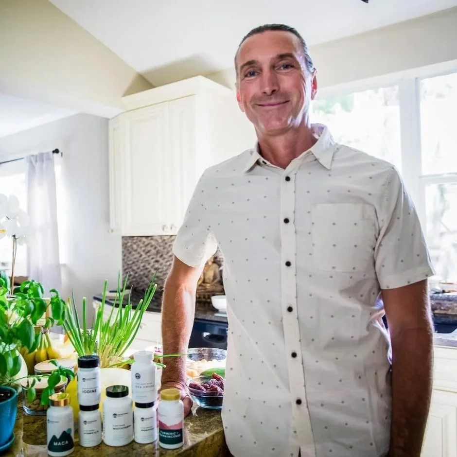 A smiling man in a white, short-sleeve, button-up shirt with small dark polka dots stands in a bright kitchen, leaning on a kitchen island with various supplement bottles, green plants, and a bowl of fruit.
