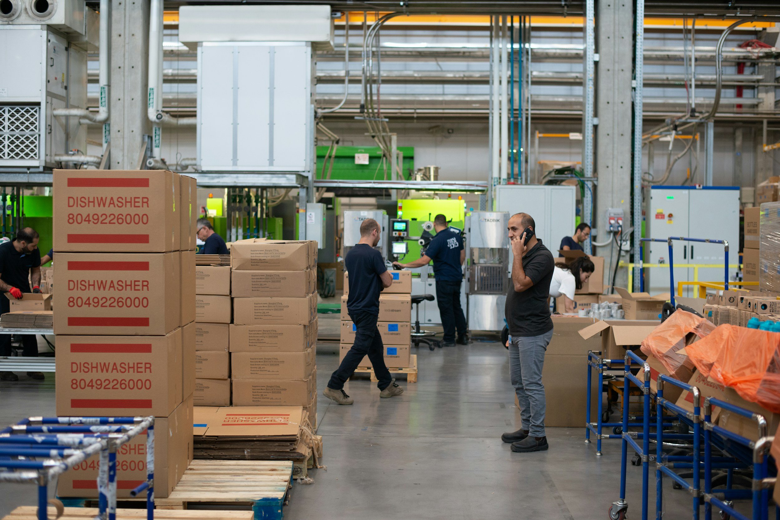 Workers in a warehouse surrounded by stacks of boxes, some labeled 'Dishwasher' with a phone number, and equipment for packaging and shipping.