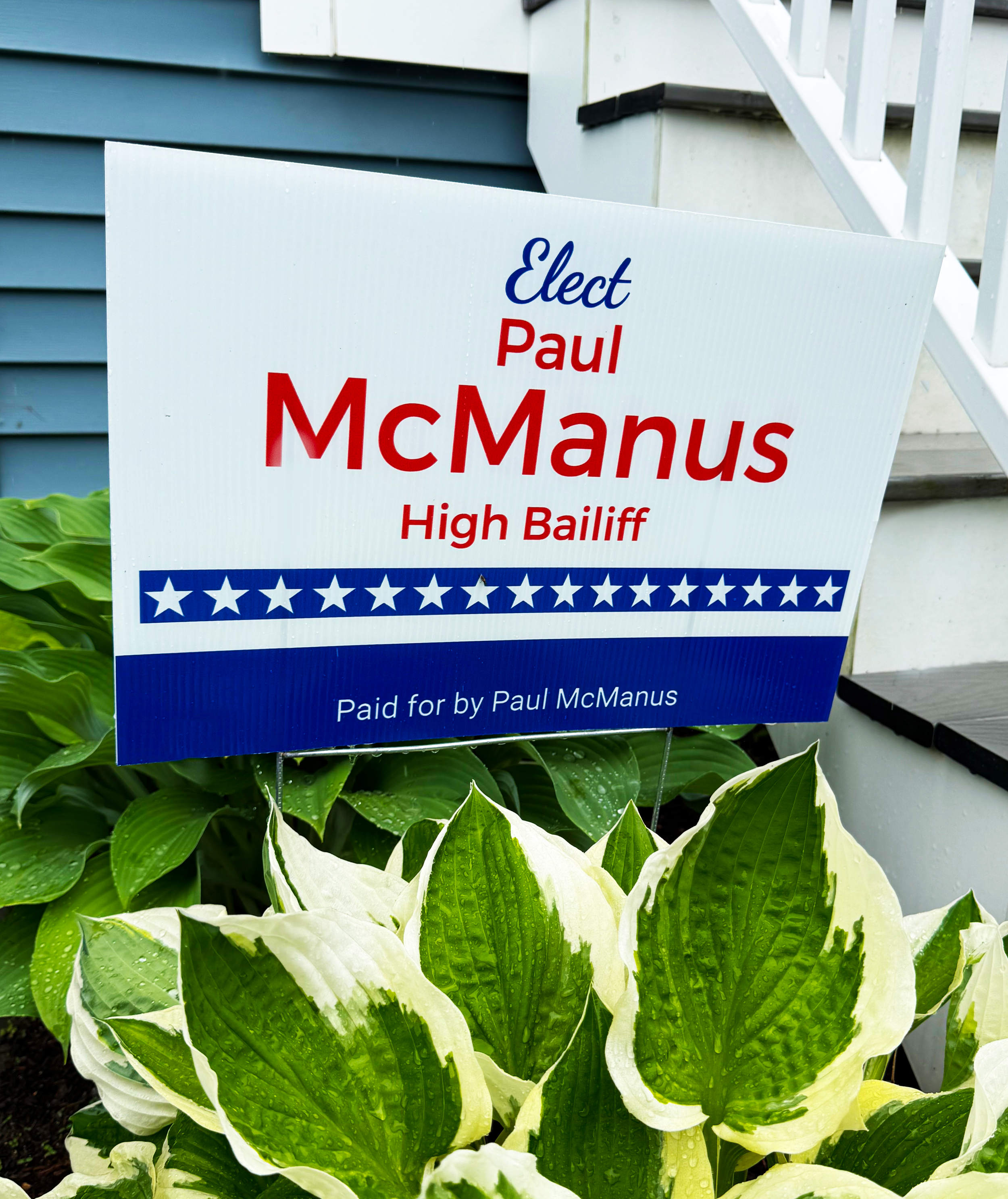 A political campaign sign in front of green leafy plants, with a house in the background. The sign reads 'Elect Paul McManus High Bailiff' and has a blue and red design with stars.