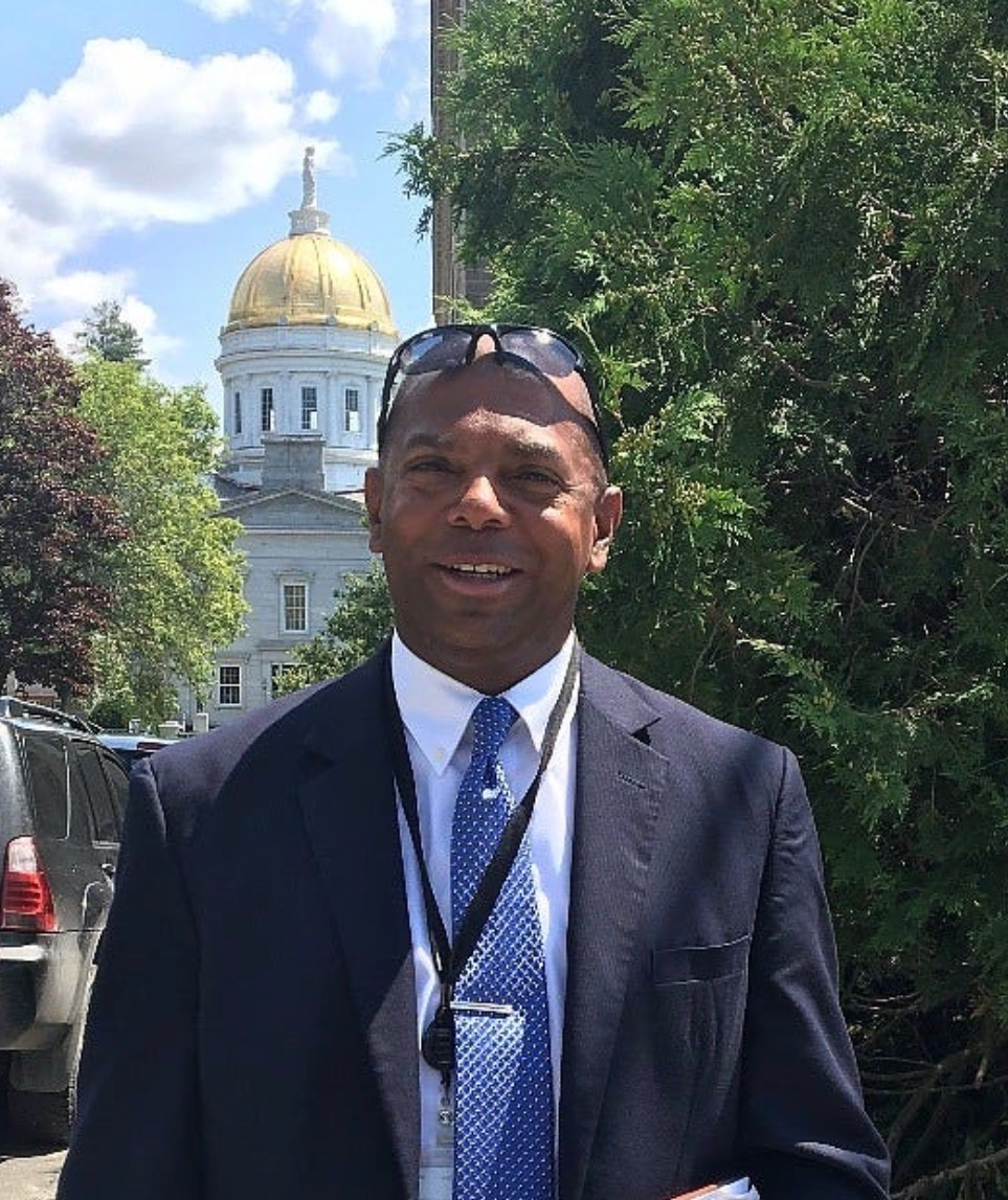 A man dressed in a dark suit, white shirt, and blue patterned tie standing outdoors in front of a leafy green background with a white and gold-domed government building behind him. He has sunglasses resting on his head and is smiling.