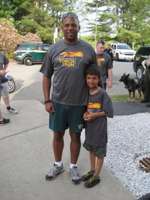 An adult man and a young boy standing outdoors, both wearing matching dark gray T-shirts with a torch and sunset design for a torch run event. The man is smiling and has short hair, while the boy is also smiling and has short dark hair. There are trees, parked cars, and other people and a dog in the background.