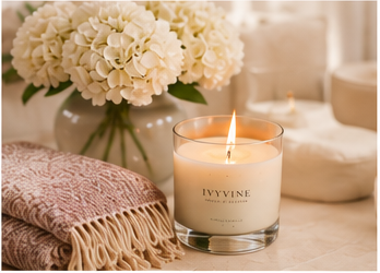 Decorative setting with a lit candle in a glass container labeled 'IyyVine', a folded pink blanket, and a bouquet of beige flowers in a vase, all on a table.