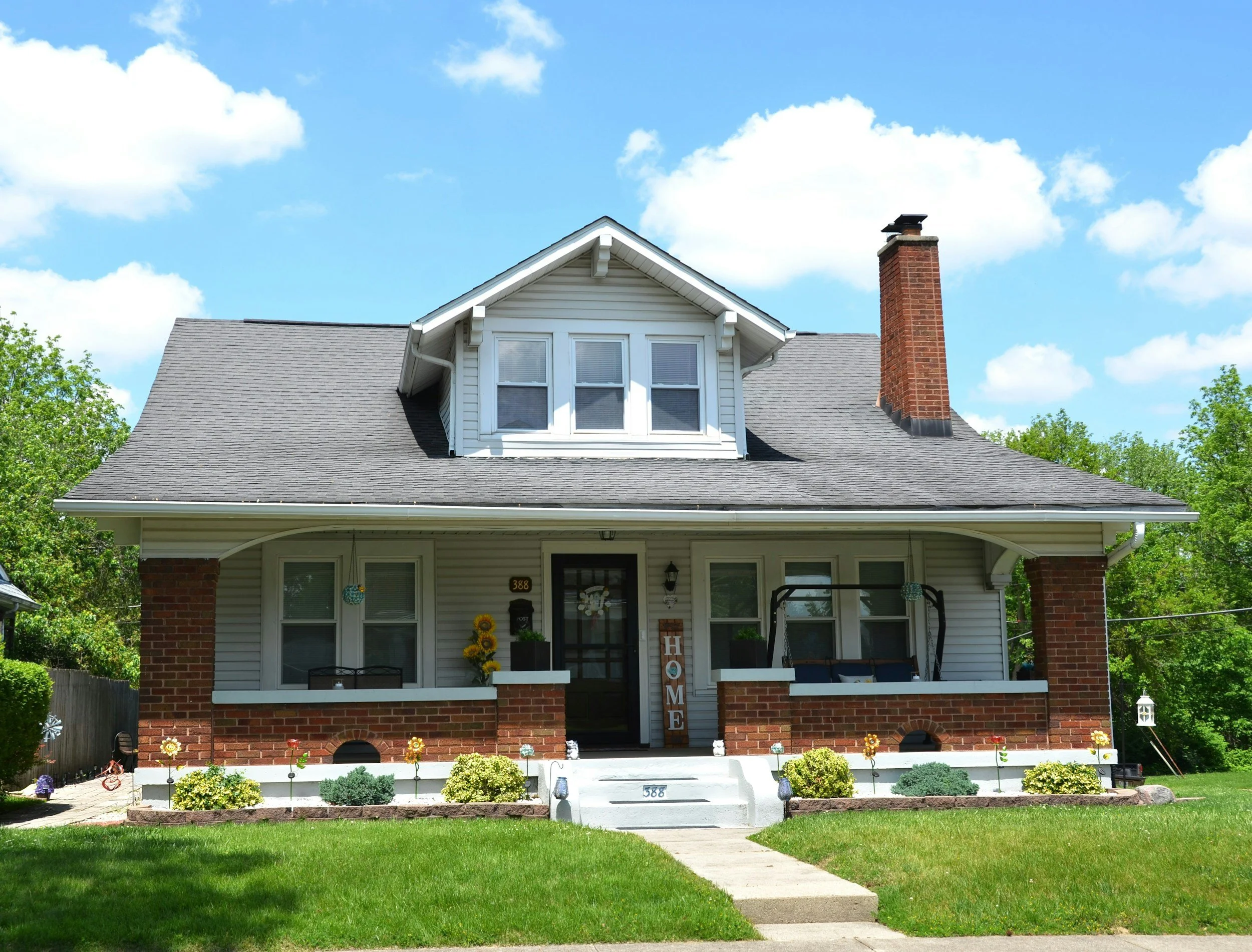 Front view of a two-story house with a front porch, brick foundation, white siding, and a gabled roof with dormer windows. There is a chimney on the right side and a green lawn with flower beds in front.
