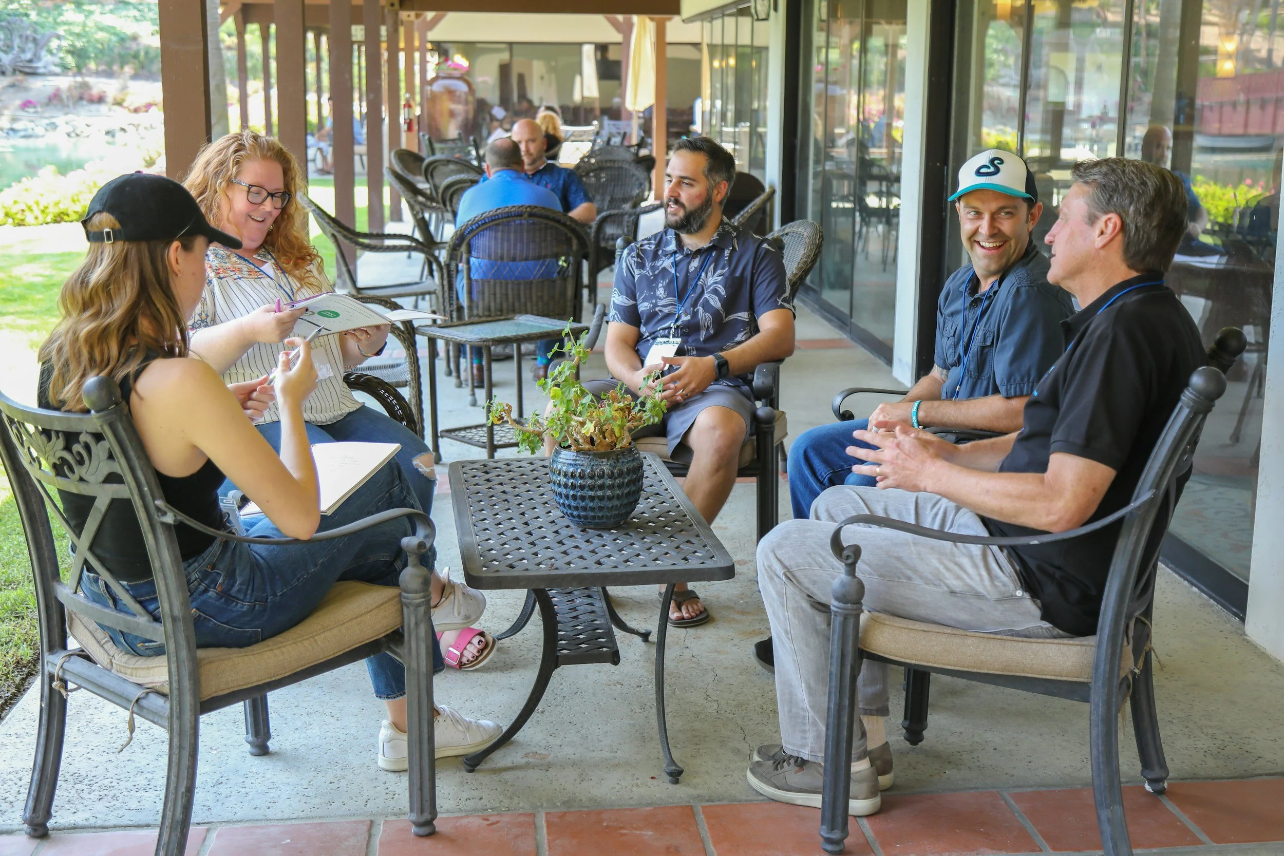 Group of five people sitting around a table outside on a patio, engaged in conversation and looking at a notebook or menu. The setting appears casual and the background shows more patio seating and greenery.