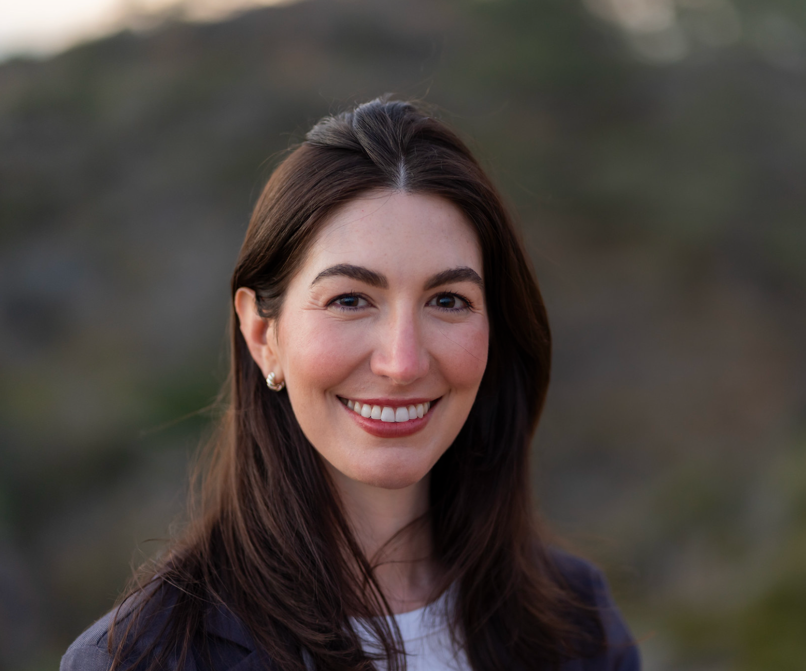 A smiling woman with brown hair, wearing earrings, outdoors in natural light.