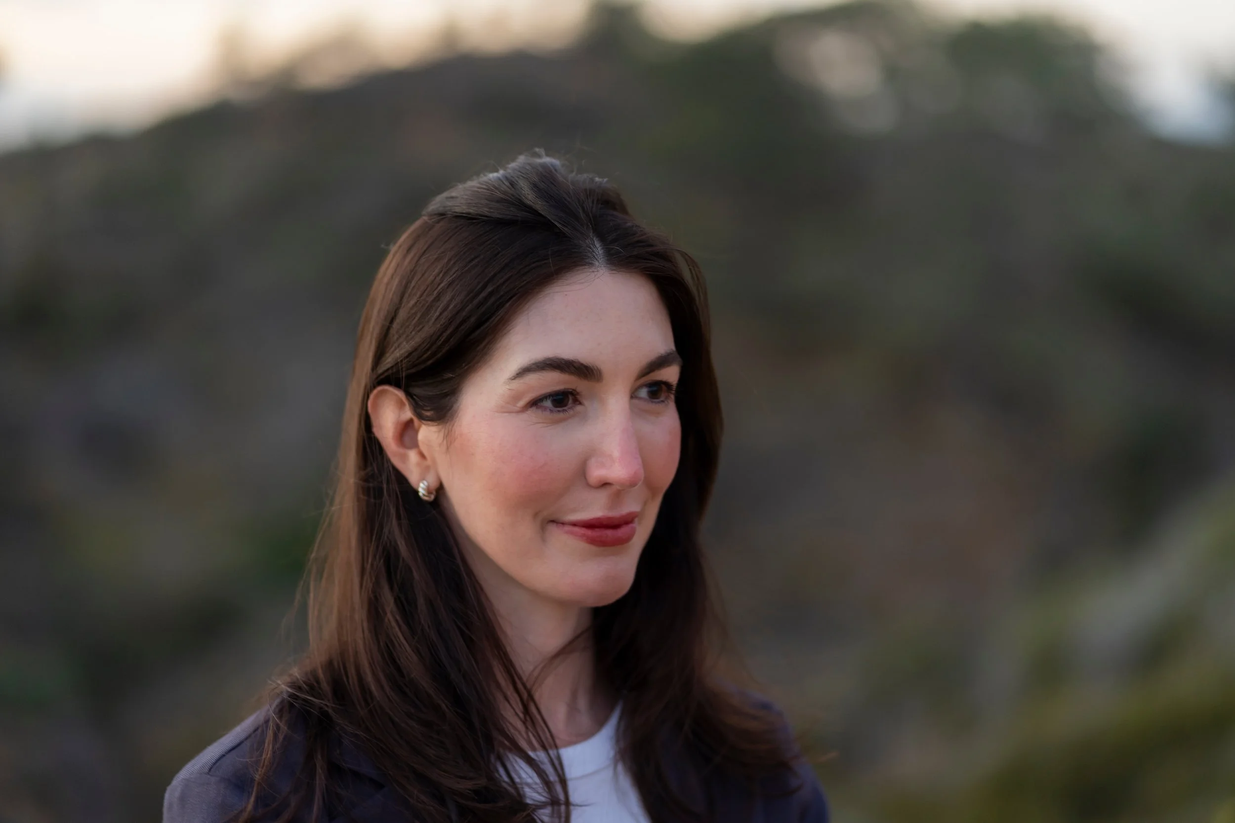 Close-up of a woman with long dark hair, wearing earrings, smiling softly with a blurred outdoor background at sunset or early evening.