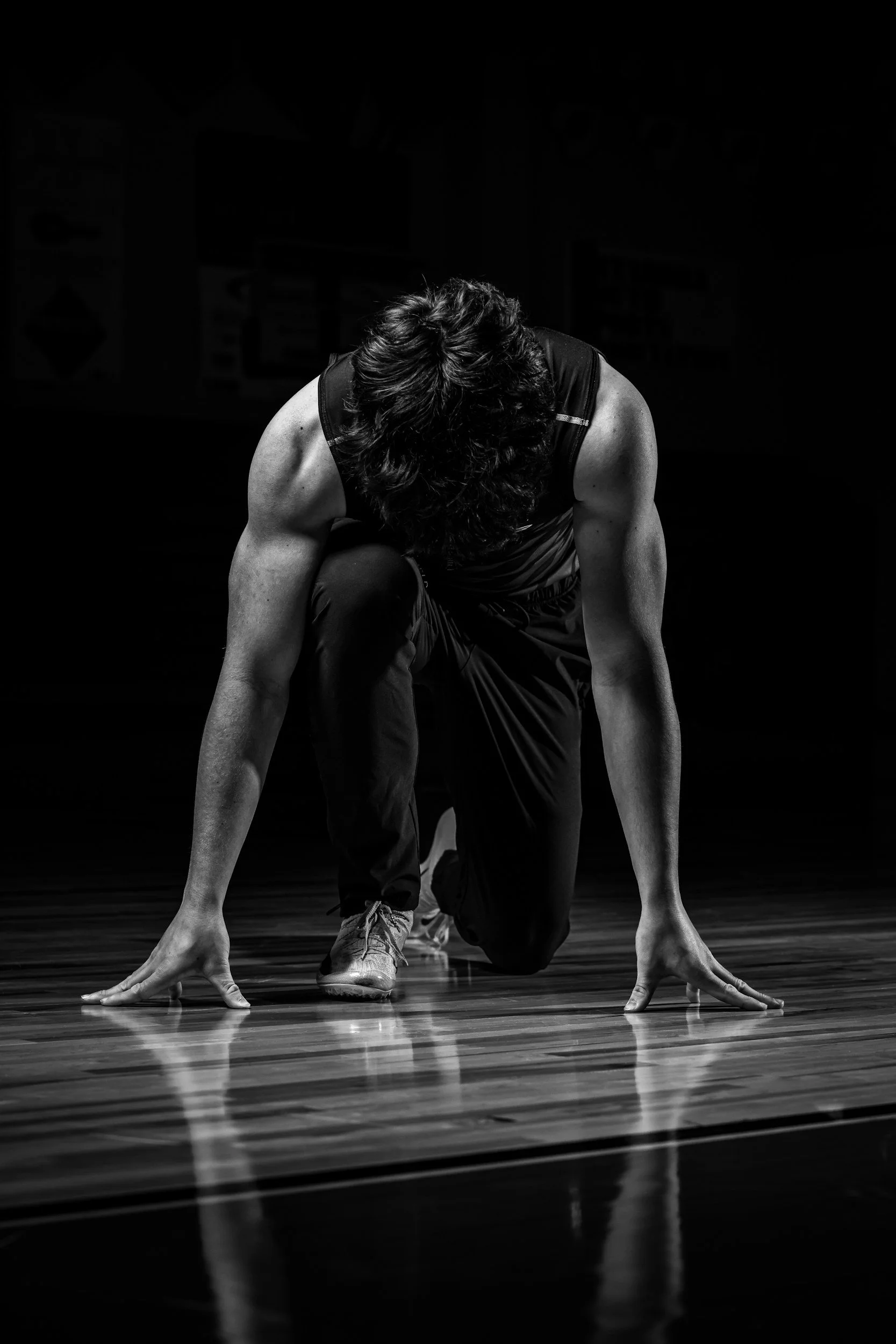 A person with curly hair performing a dance move on a dark stage, crouched on one knee with hands touching the floor, wearing athletic clothing.