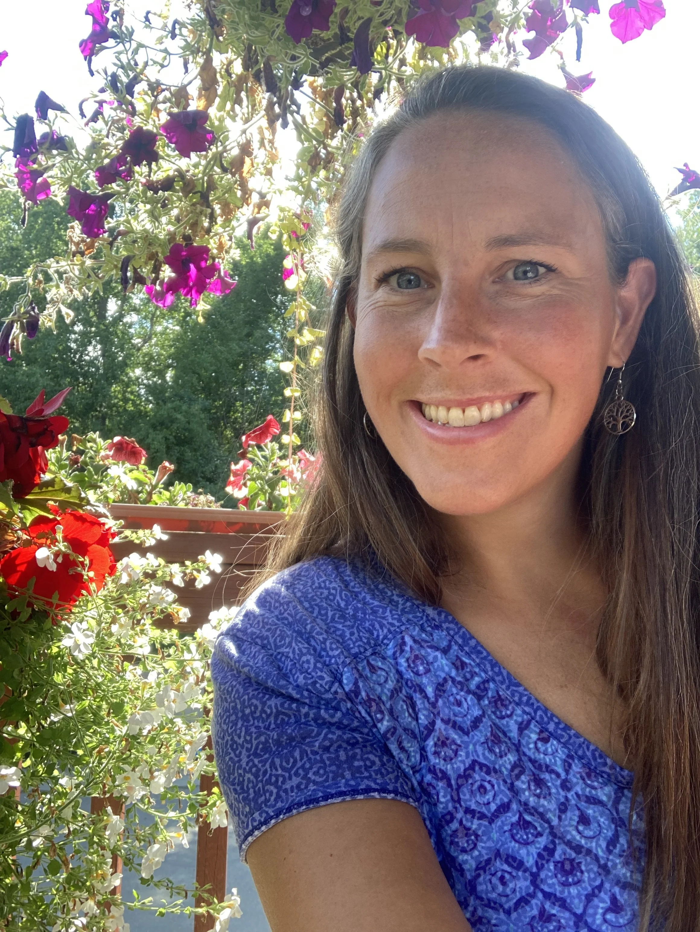 A woman smiling outdoors with a background of flowering plants, including purple and red flowers and green trees, wearing a blue patterned shirt and a tree leaf earring.