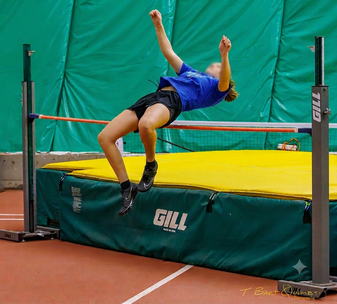 An athlete in a blue shirt and black shorts falls over the high jump bar during an indoor track event.
