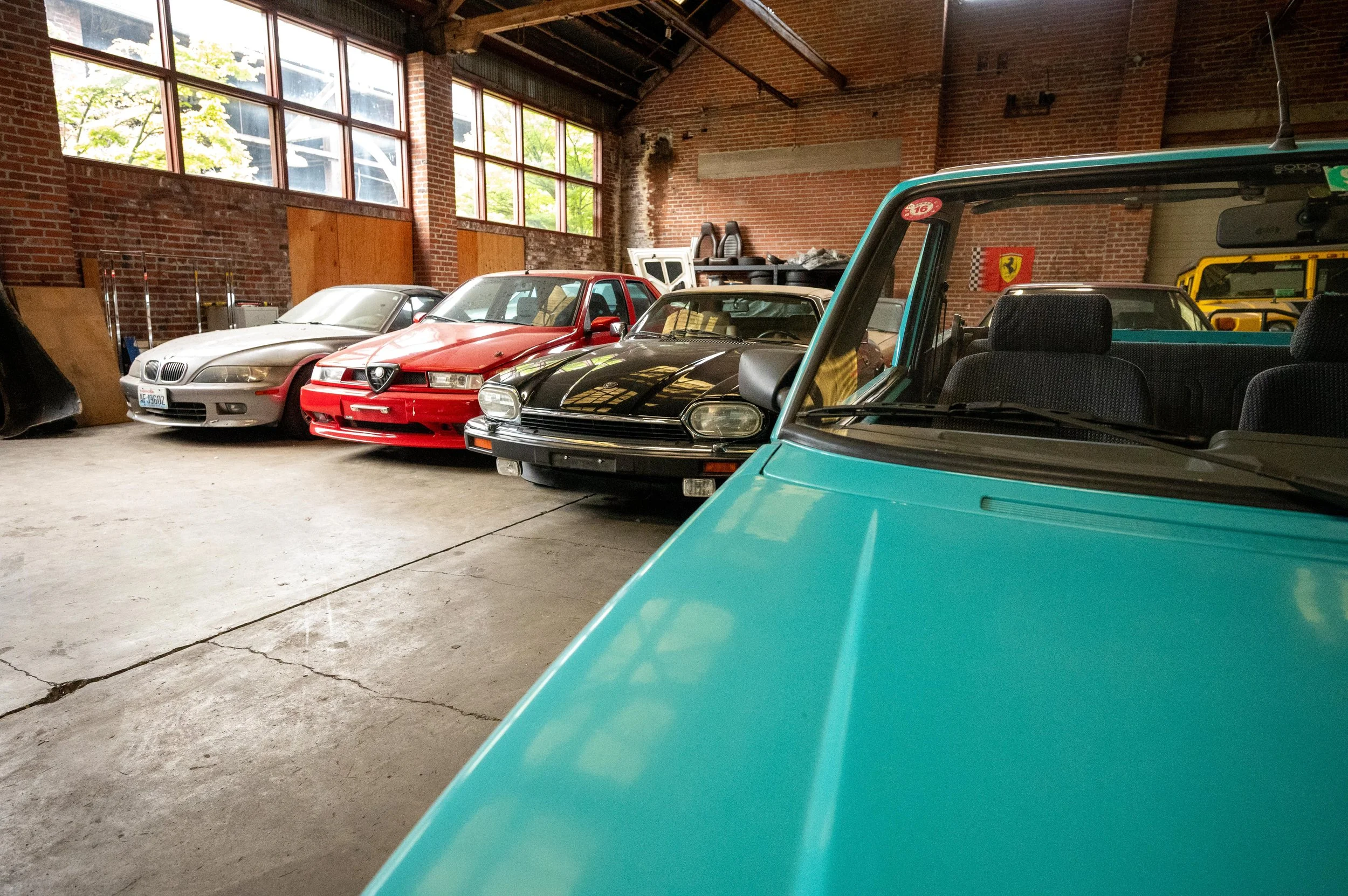 A row of classic cars inside a warehouse with brick walls and large windows, including a turquoise convertible in the foreground, followed by a black sports car, a red car, and a beige car.