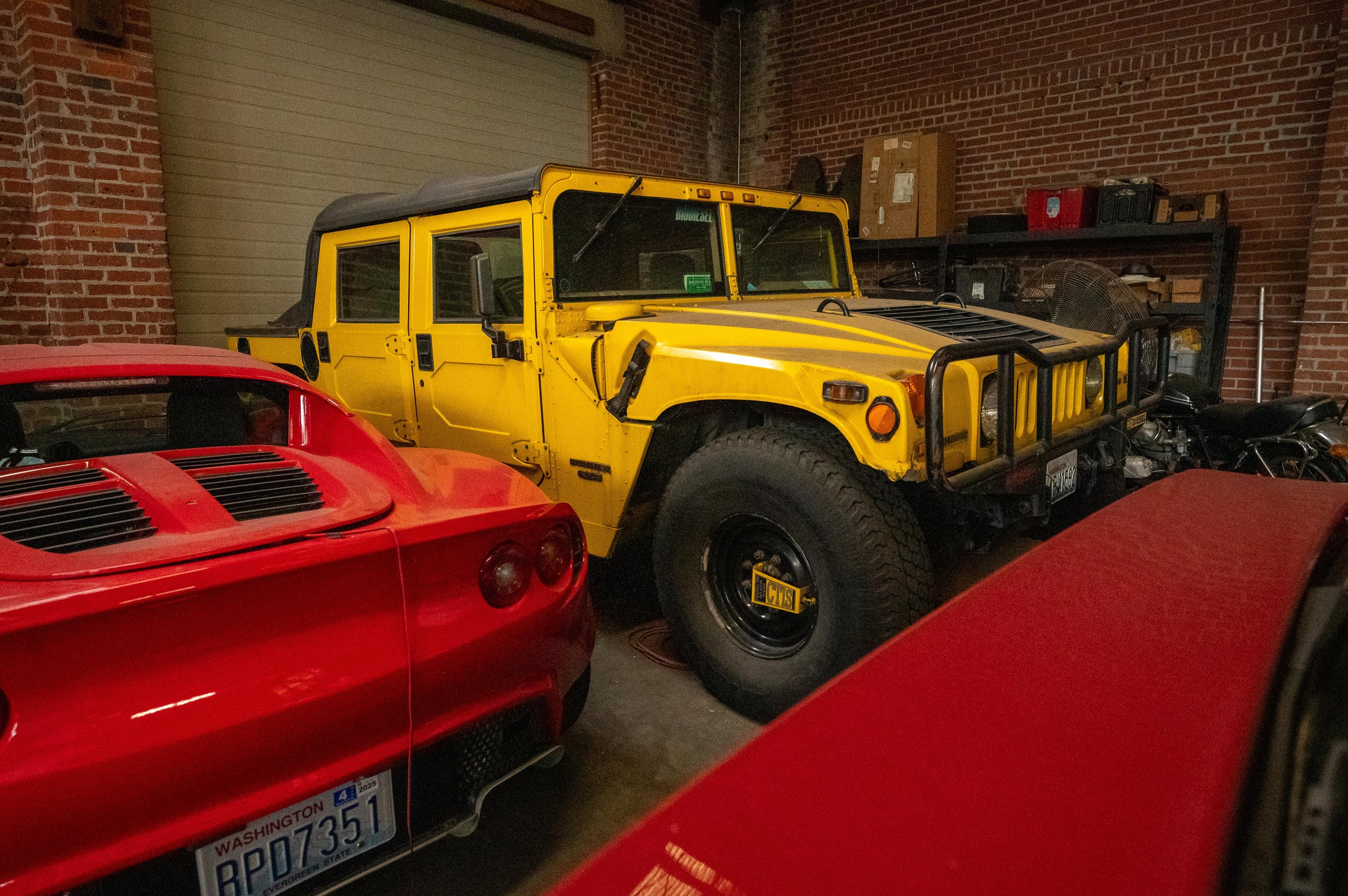 A collection of cars in a garage, including a yellow Hummer, red sports cars, and a motorcycle, with exposed brick walls and storage shelves in the background.