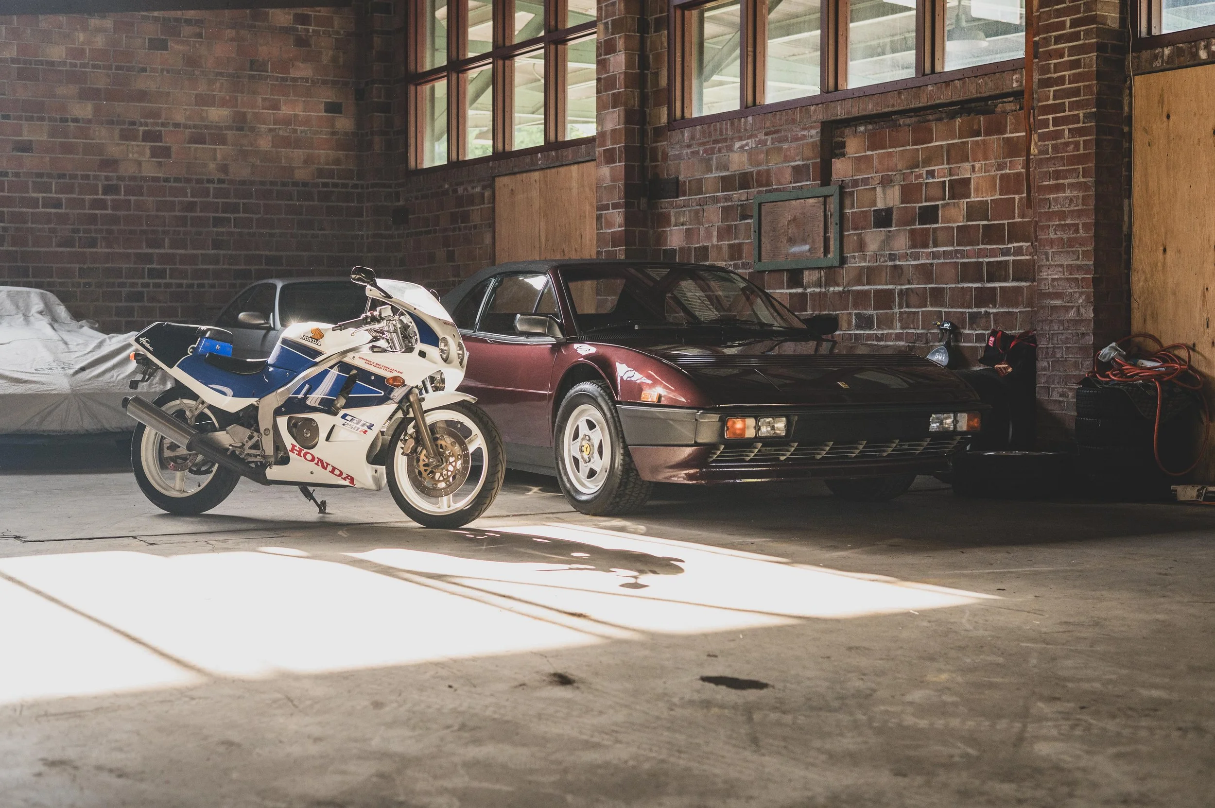 A garage with brick walls and large windows, containing a white and blue Honda motorcycle, a black sports car, and some tools and equipment underneath a window.
