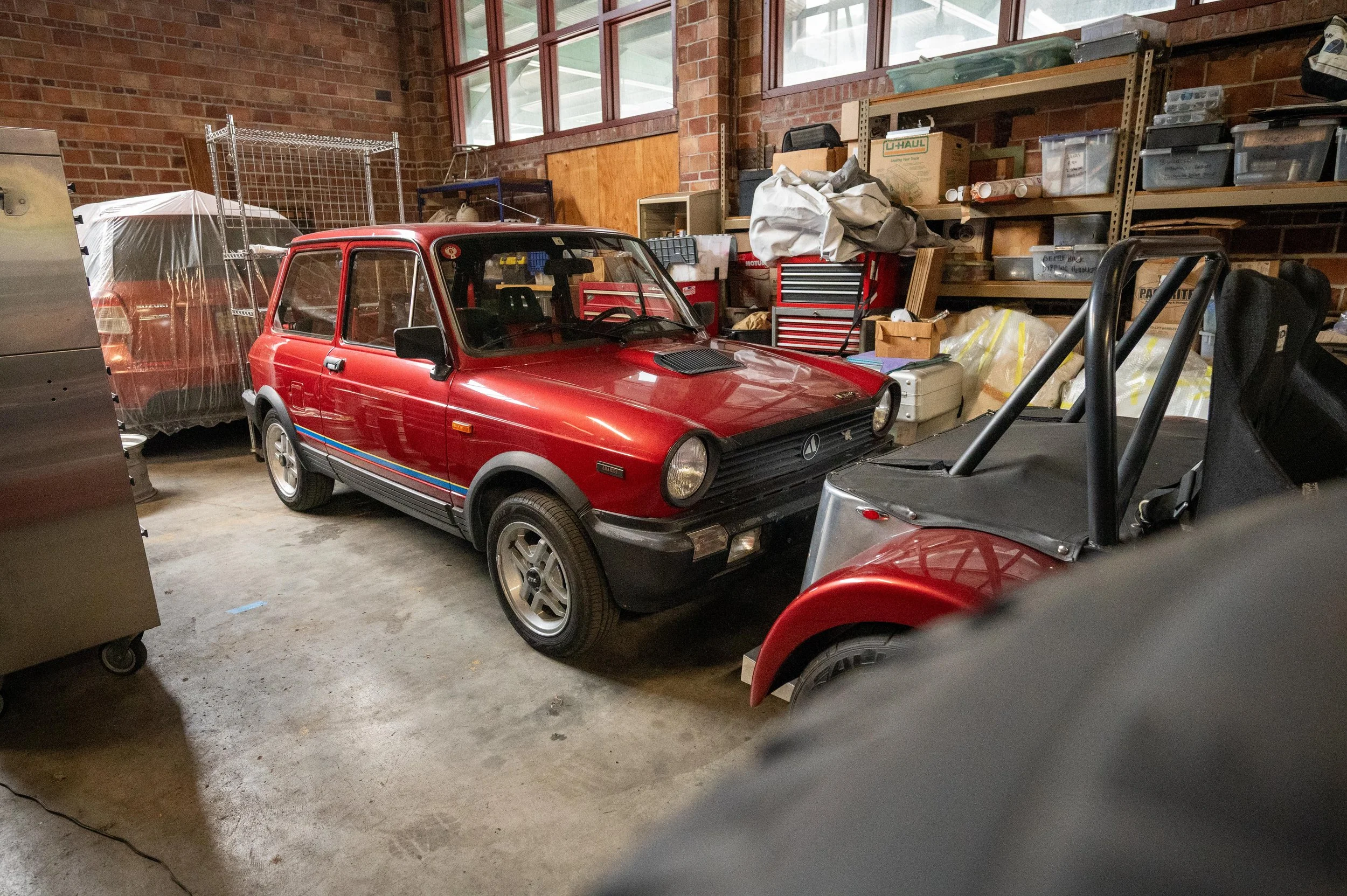 A red vintage car in a cluttered garage with shelves filled with boxes and tools.