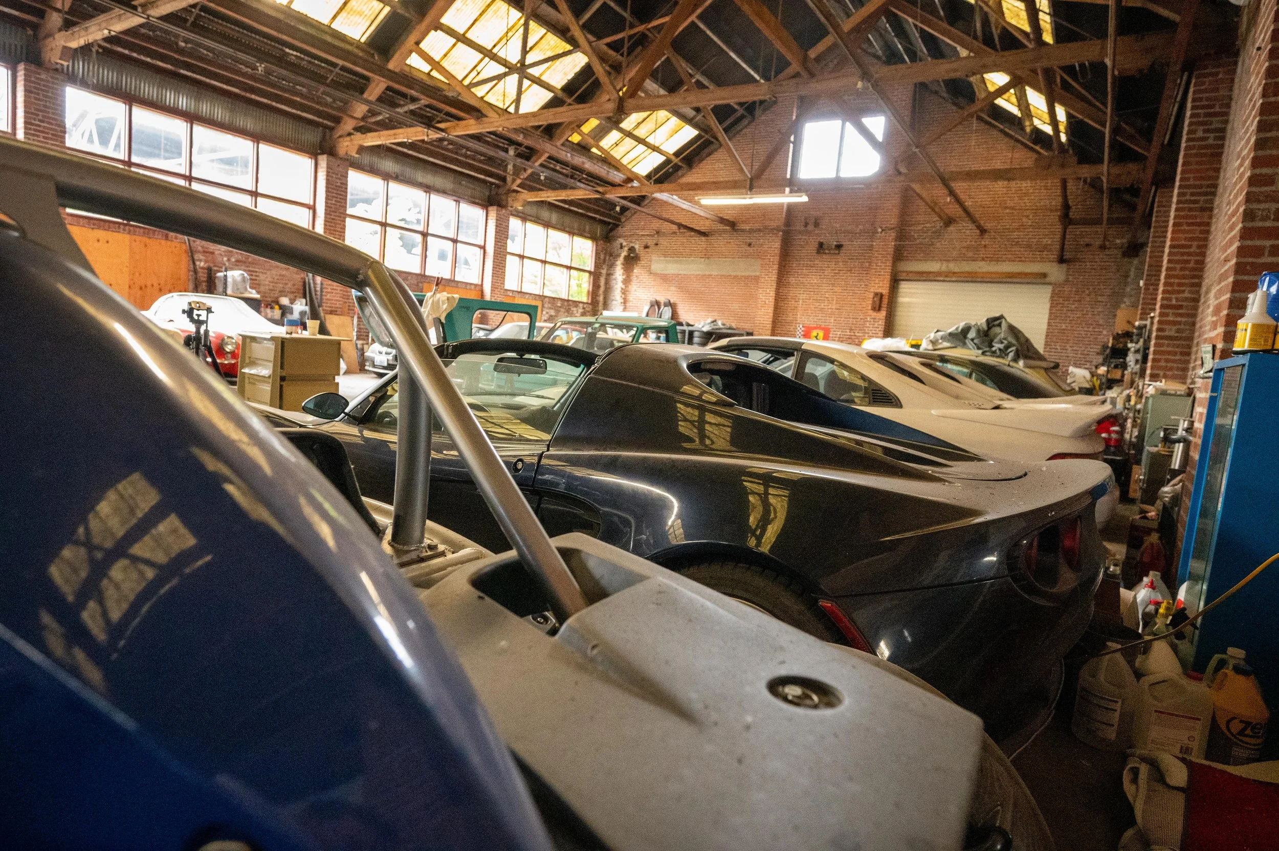 An indoor garage with several vintage and sports cars parked inside. The garage has brick walls, large windows, and a wooden ceiling with exposed beams. Various tools and supplies are visible around the space.
