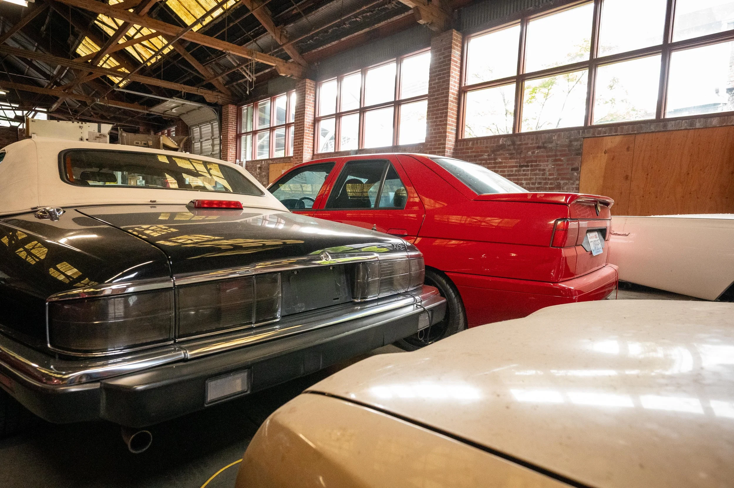 Interior of a garage with several vintage cars parked inside, featuring brick walls and large windows letting in natural light.