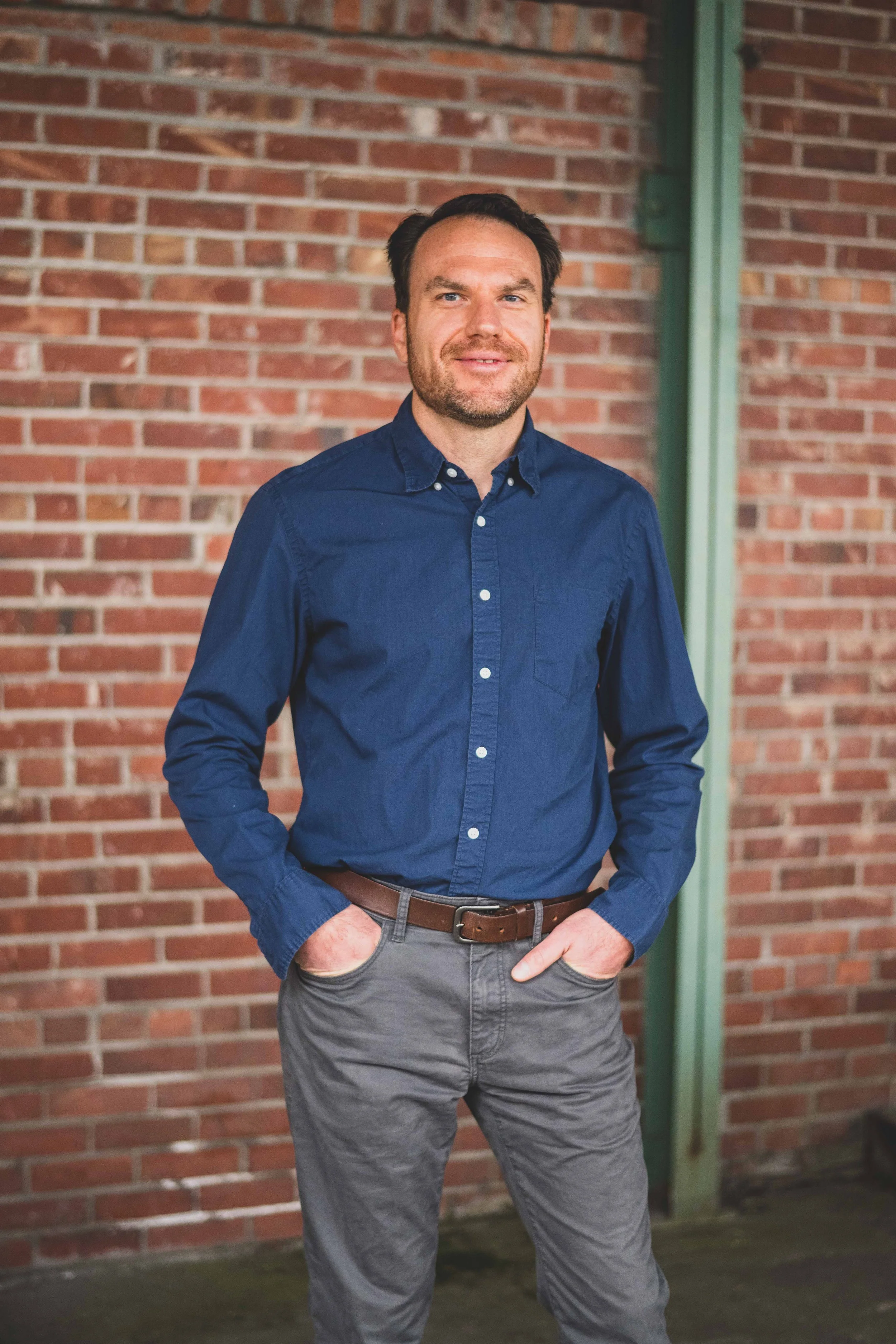 A man standing outdoors in front of a brick wall, wearing a navy blue button-up shirt and gray pants, with one hand in his pocket and looking at the camera.