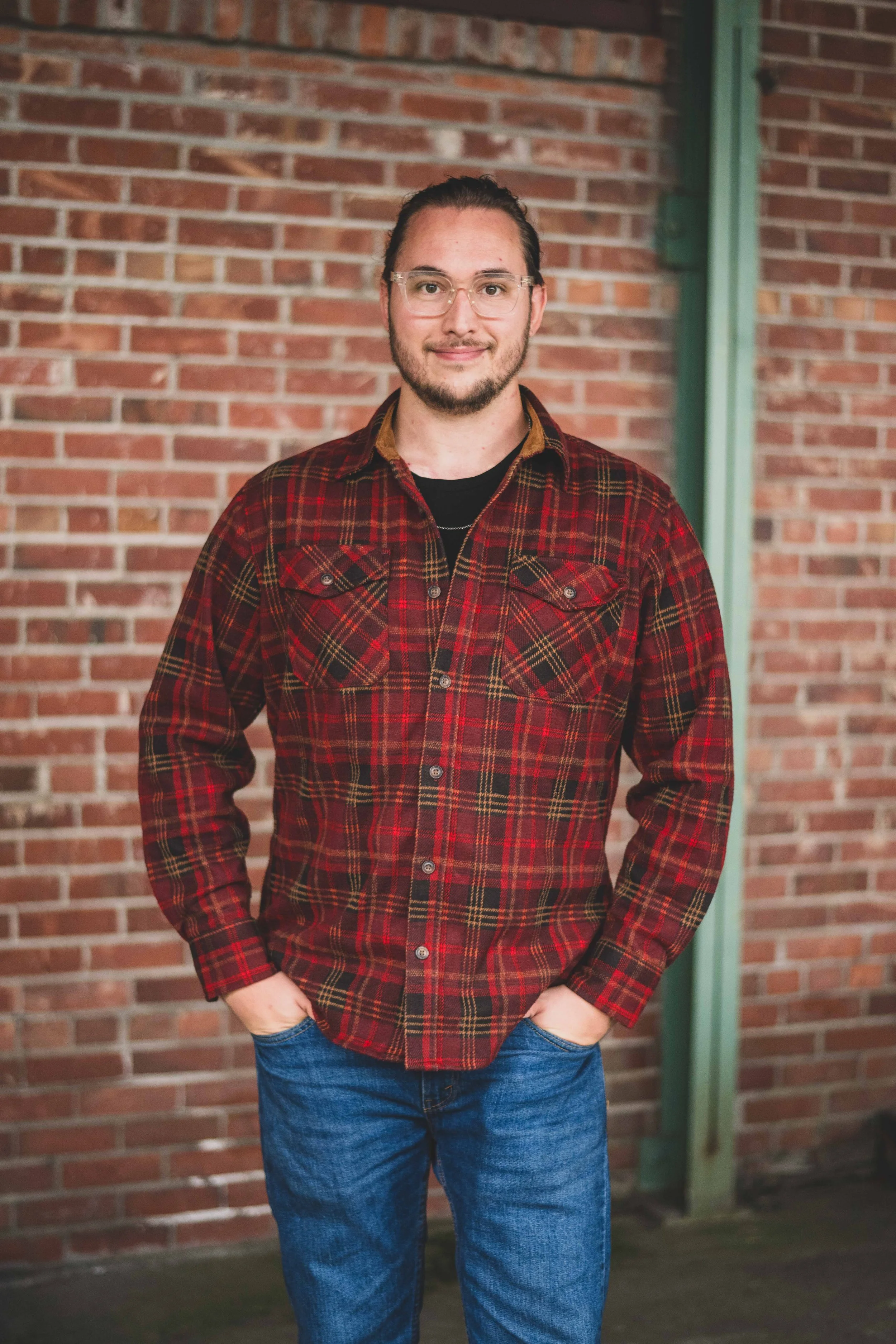 A young man with glasses, a beard, and long hair tied back, wearing a red plaid shirt over a black T-shirt and blue jeans, standing with hands in pockets in front of a brick wall.