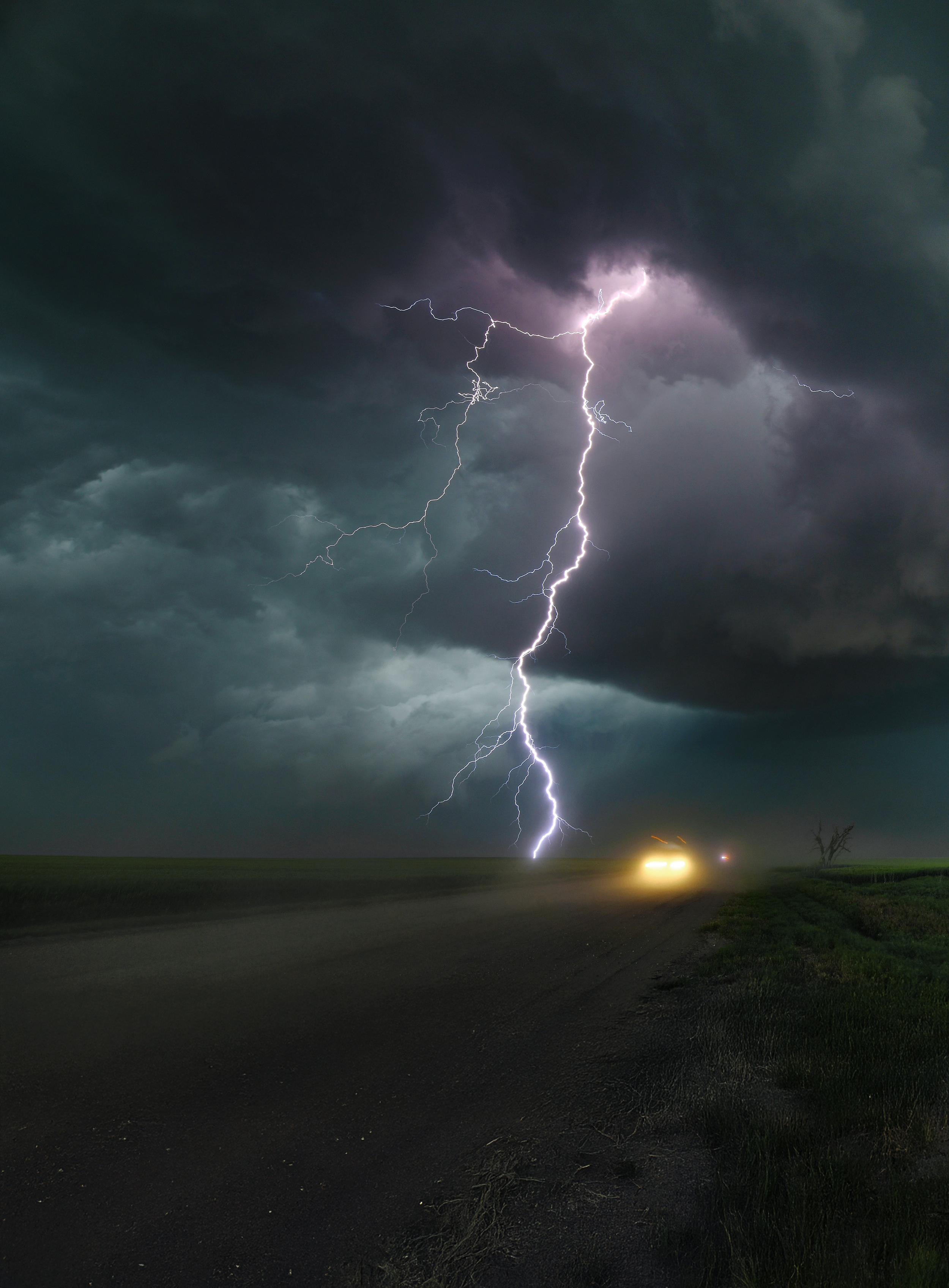 Lightning strikes in a stormy night sky over a rural road, with car headlights visible in the distance and a leafless tree on the right.