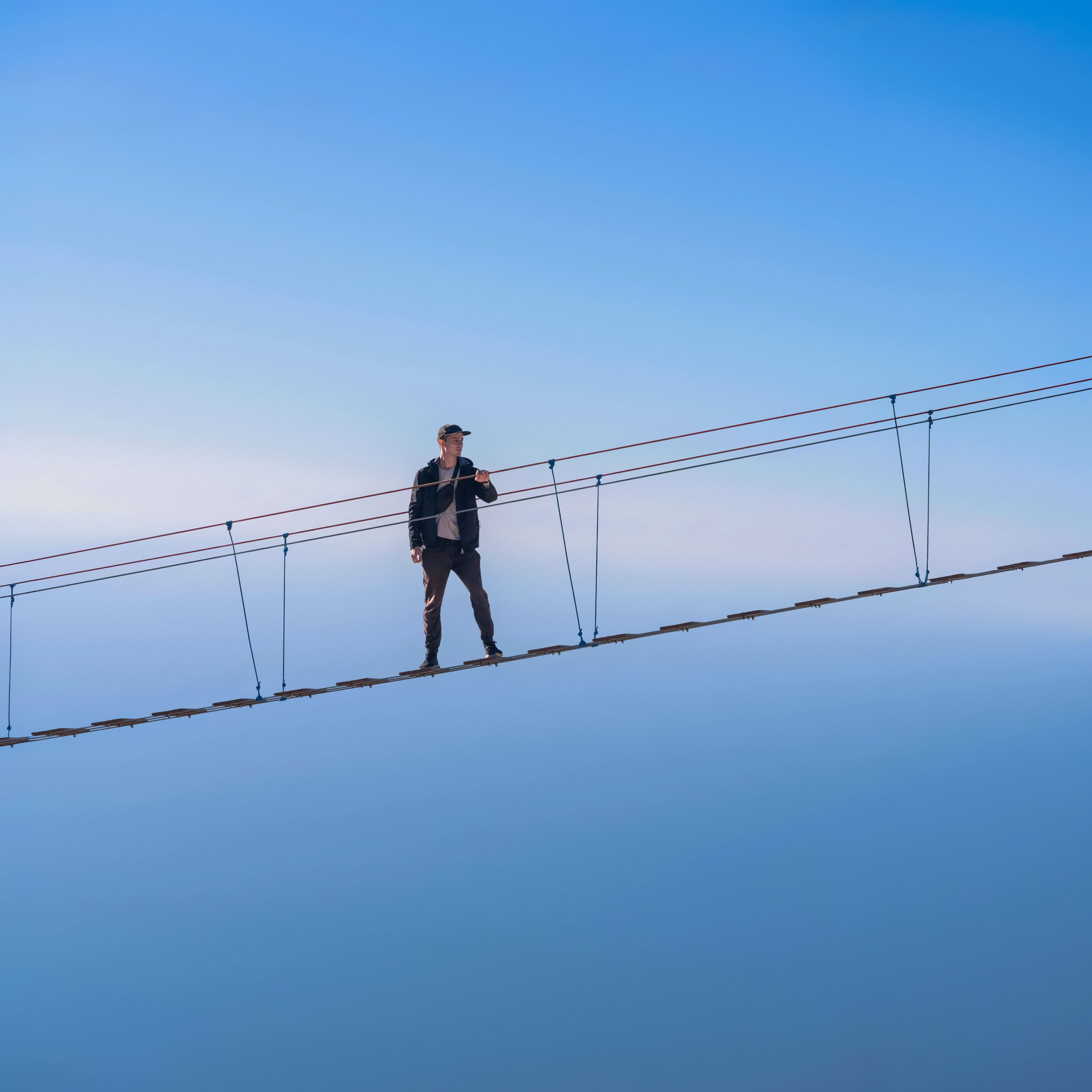 A person wearing a cap and dark clothing walking on a suspension bridge with cables against a bright blue sky.