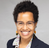 Close-up portrait of a smiling woman with short curly hair, wearing glasses, earrings, and a patterned shirt.