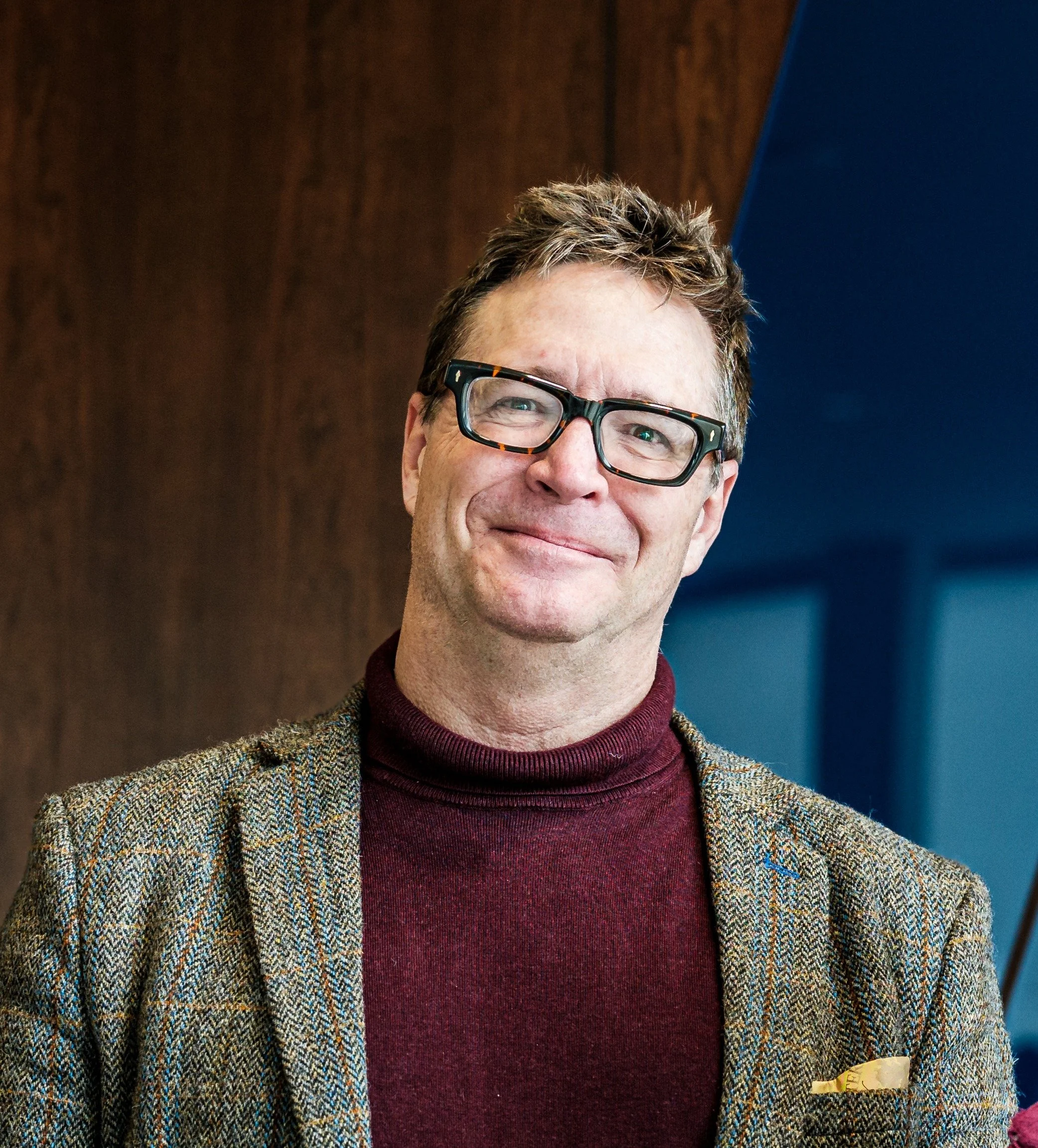 Close-up of smiling man wearing glasses, a maroon turtleneck, a tweed blazer, and a yellow pocket square, standing against a wooden and blue background.