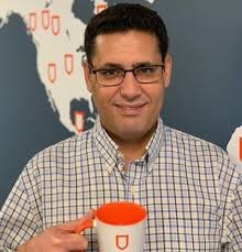 Man with glasses and dark hair holding a white mug with an orange interior, standing in front of a world map background.