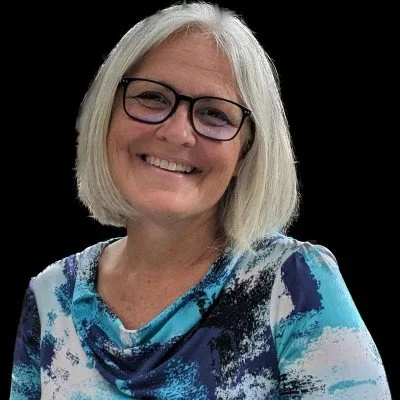 A smiling woman with shoulder-length white hair, wearing glasses and a blue, white, and black patterned top, against a black background.