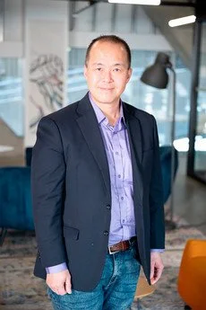 Professional man wearing a dark blazer and light blue shirt standing in a modern office.