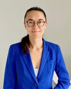 A woman with glasses and a ponytail wearing a blue blazer and white top, standing against a plain light background.