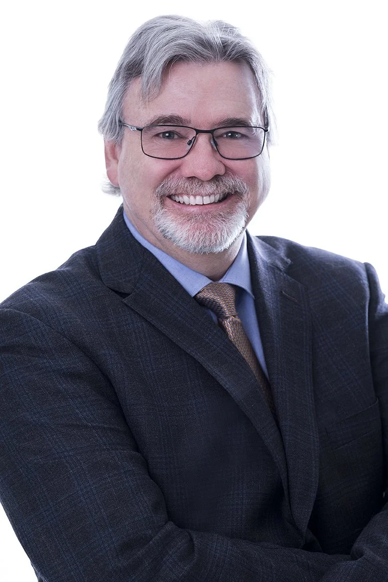 A smiling middle-aged man with gray hair, beard, and glasses, wearing a dark suit, light blue shirt, and patterned tie, against a white background.