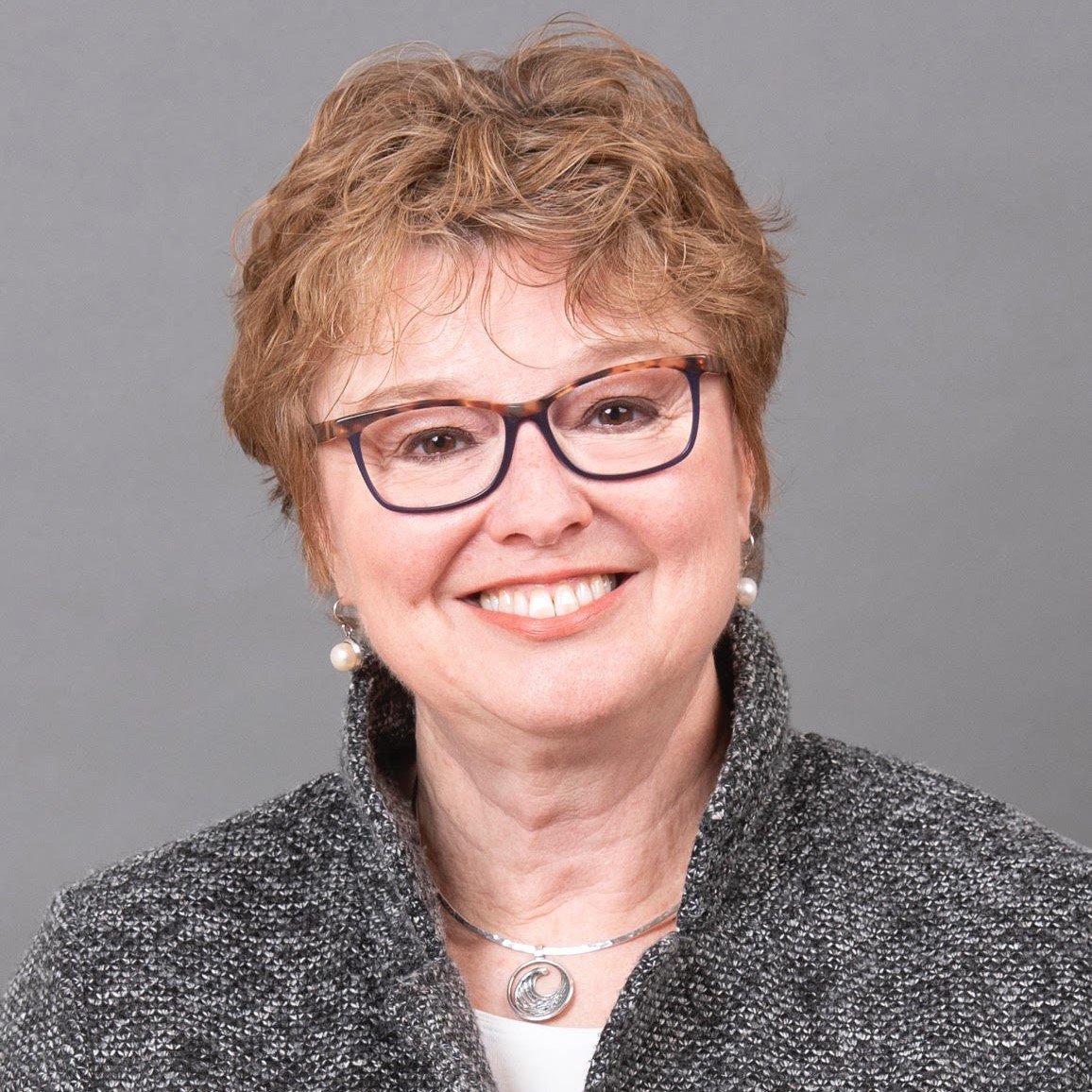 A smiling middle-aged woman with short, curly red hair, wearing glasses, pearl earrings, a silver necklace with a pendant, and a textured gray jacket against a plain gray background.
