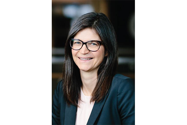 A woman with shoulder-length dark hair, wearing glasses, a navy blazer, and a white top, smiling at the camera in an indoor setting.