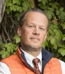 Portrait of a man with light hair and beard, wearing a white shirt, orange vest, and tie, standing outdoors with green leaves in the background.