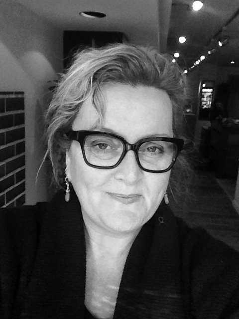 A black and white photo of a woman with wavy short hair wearing glasses and earrings, smiling slightly, in an indoor setting.