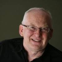 A smiling middle-aged man with glasses and short hair, wearing a black shirt, against a dark background.