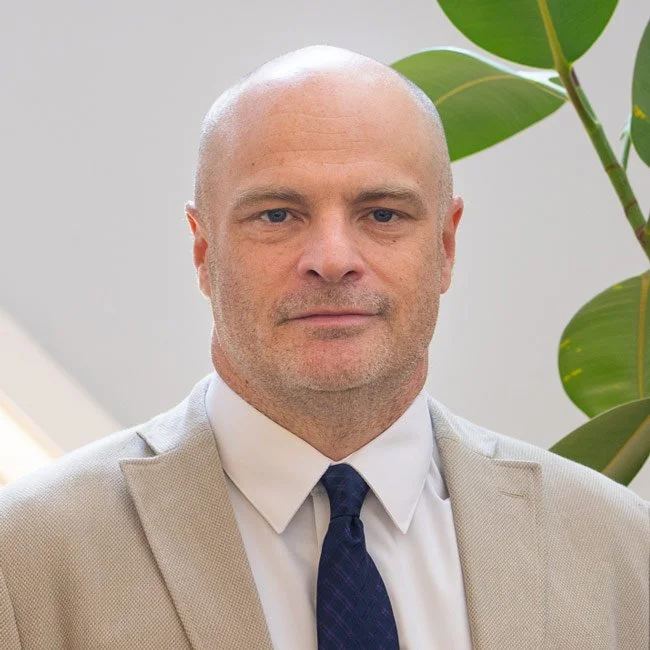 A bald man in a light gray suit, white shirt, and dark blue tie, standing indoors with green houseplants in the background.