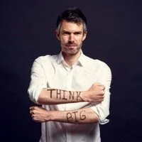 Man with arms crossed, wearing a white shirt, with the word 'THANK' written on his left forearm and the word 'GOD' on his right forearm.