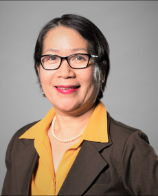Portrait of a woman wearing glasses, a brown blazer, and a yellow blouse, smiling against a gray background.