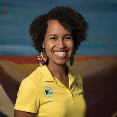 A woman smiling, wearing a yellow polo shirt with a logo, and large colorful earrings, standing in front of a painted background.