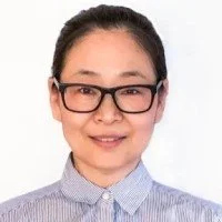 A woman with glasses and black hair in a bun, wearing a blue and white striped shirt, smiling against a white background.