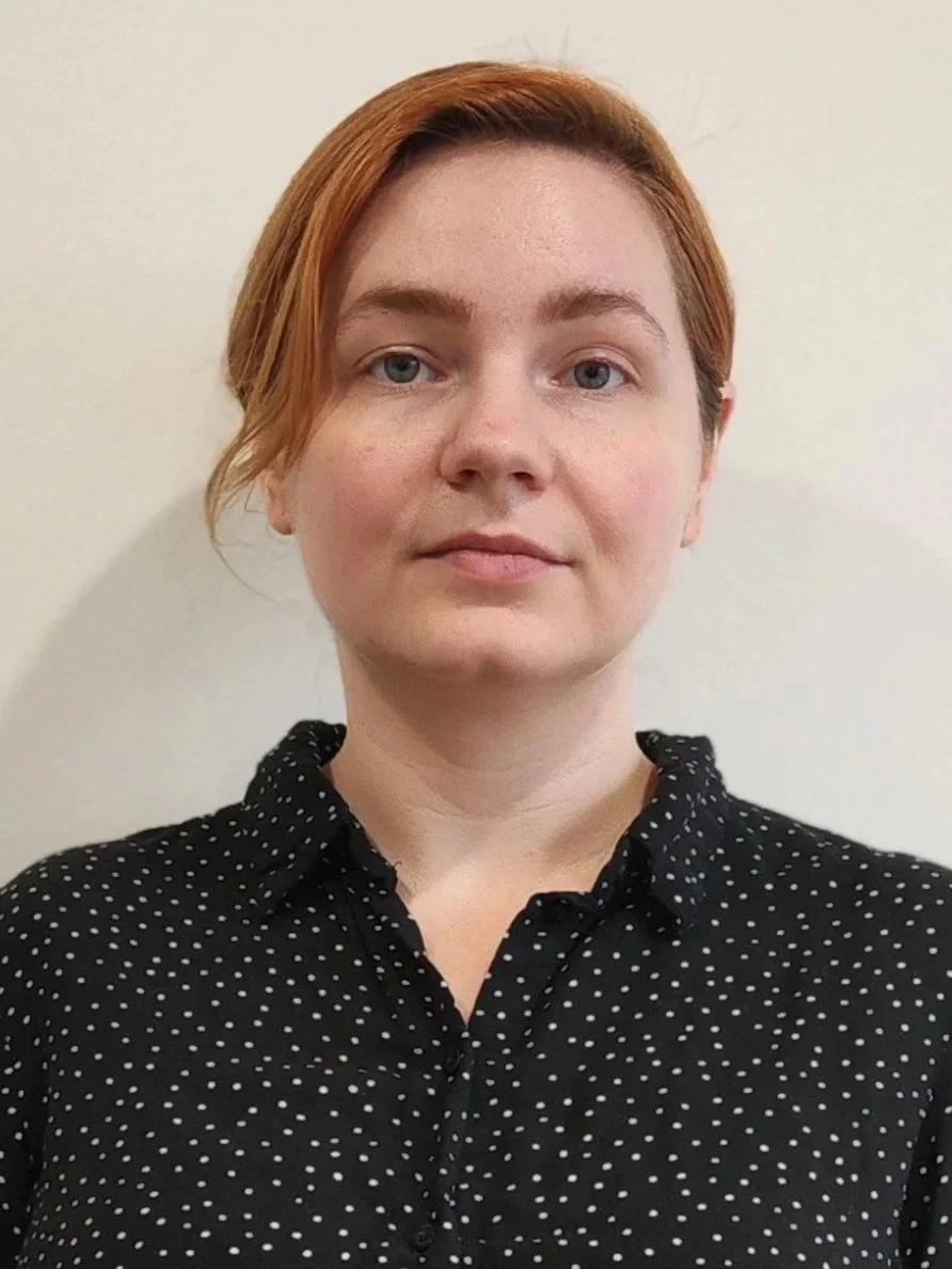 A woman with short red hair, blue eyes, wearing a black shirt with white polka dots, standing against a plain light-colored background.
