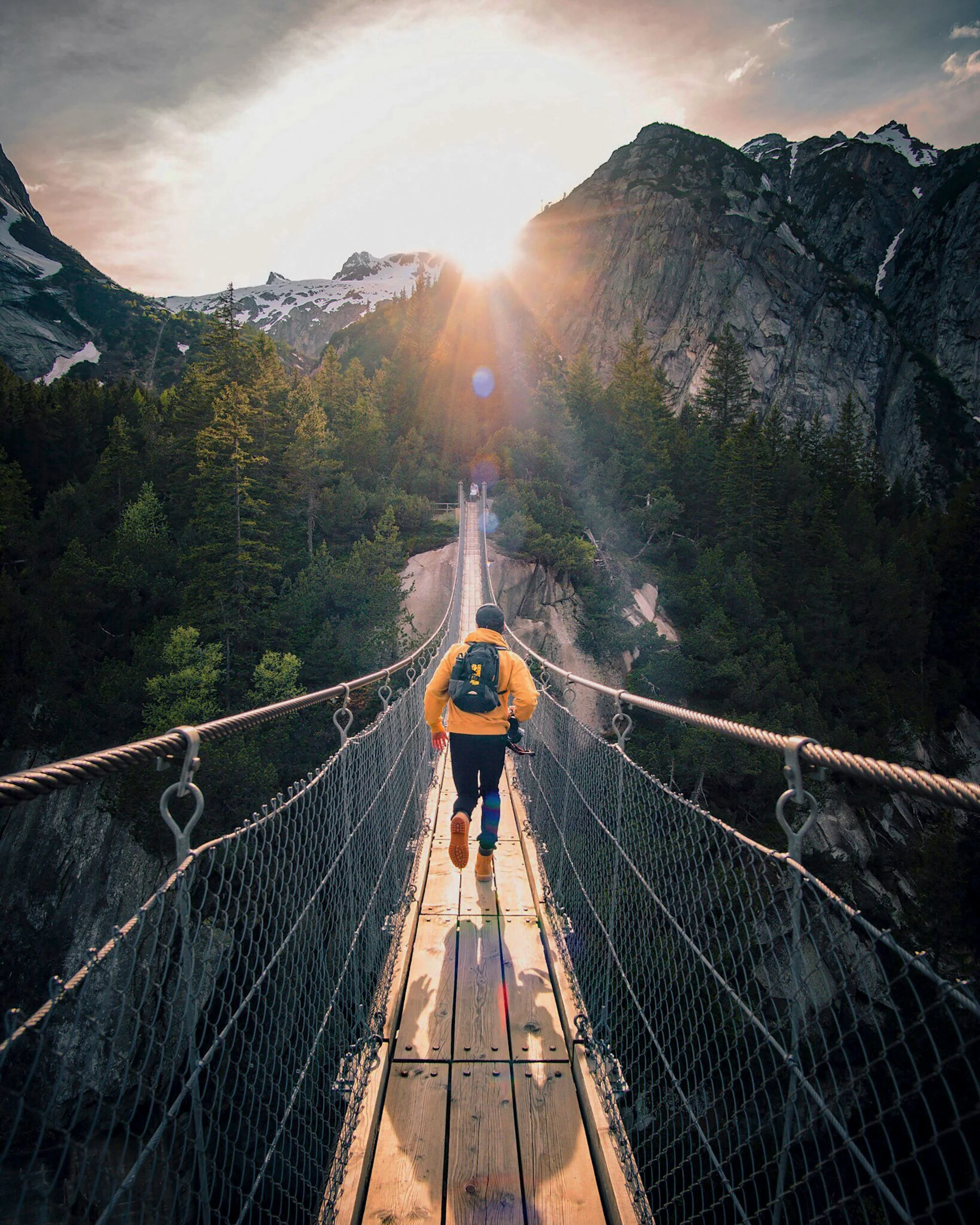 Person wearing yellow jacket and backpack walking on a suspension bridge in a mountainous area with snow-capped peaks and dense green forest, sunlight shining in the sky.