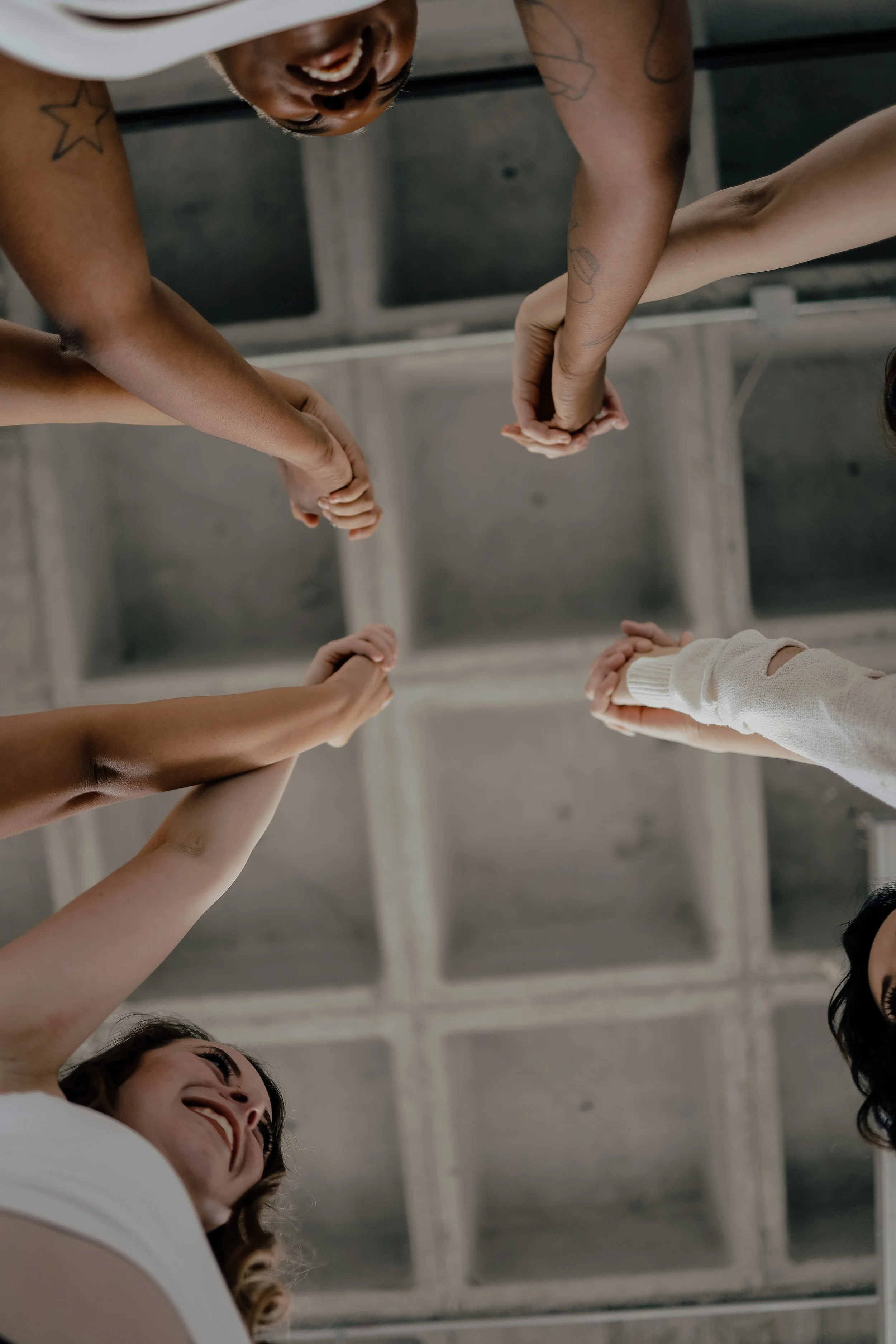 A group of diverse women holding hands in a circle, smiling, indoors with a ceiling featuring exposed beams.