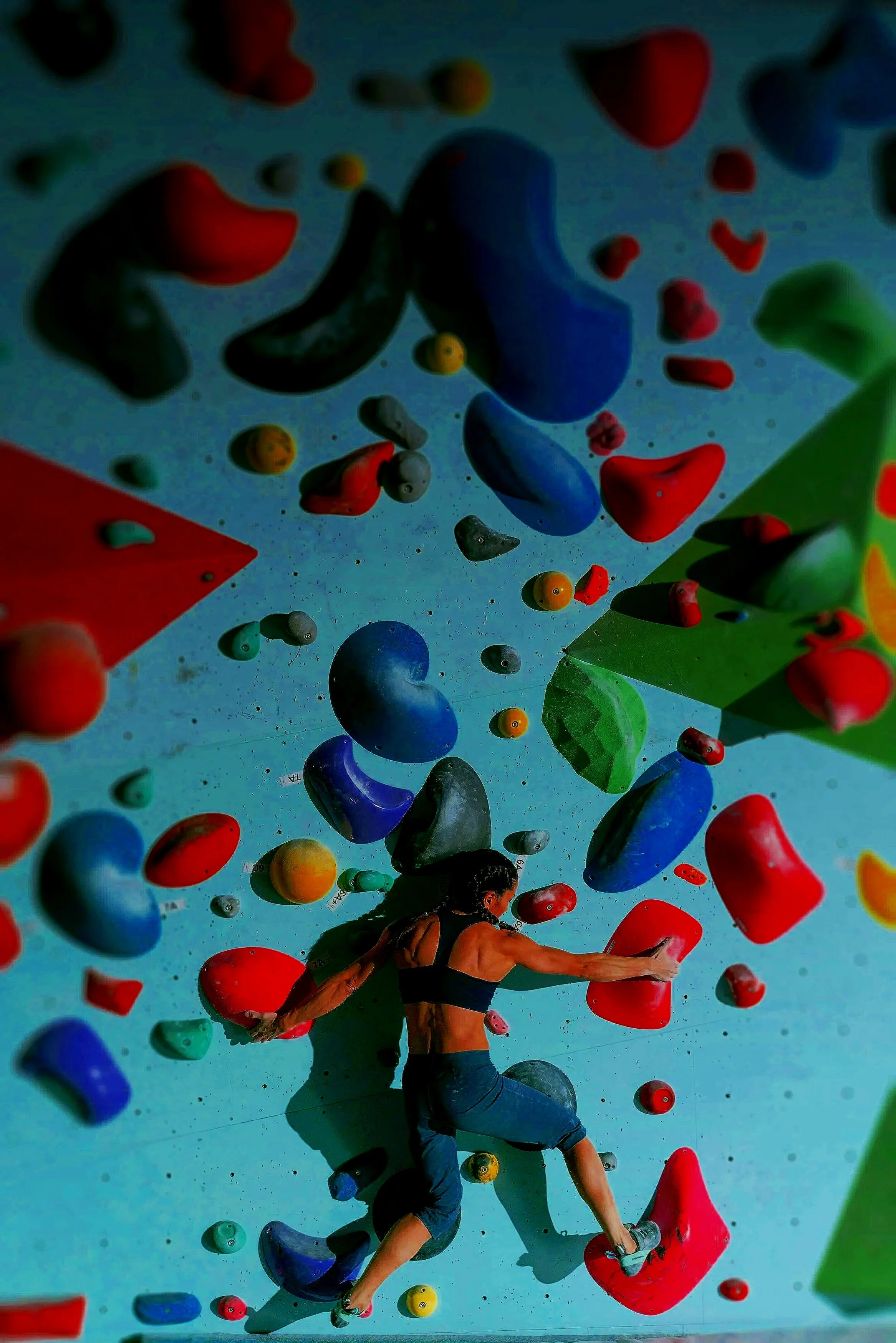 A man in a tank top and shorts bouldering on an indoor climbing wall with various colored holds.