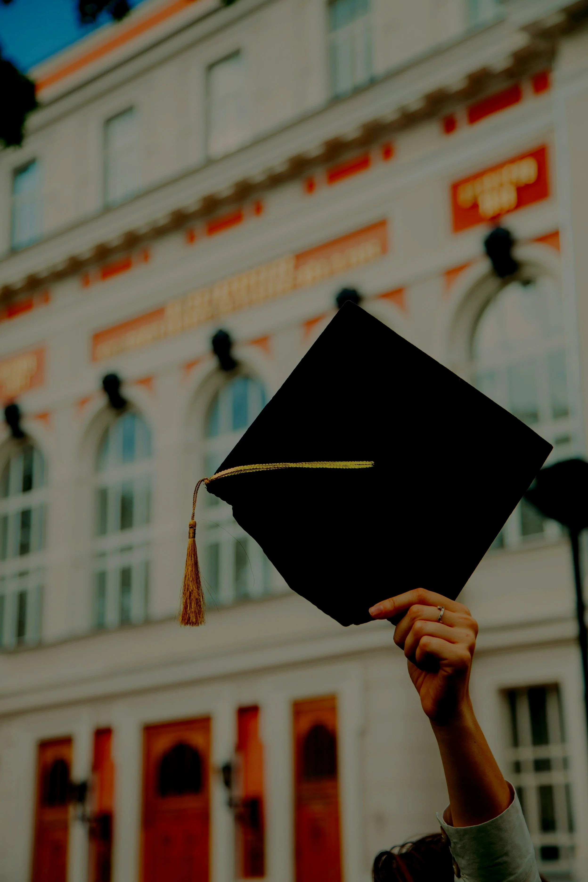 A person holding a graduation cap in front of a historic building with large windows and ornate architecture.