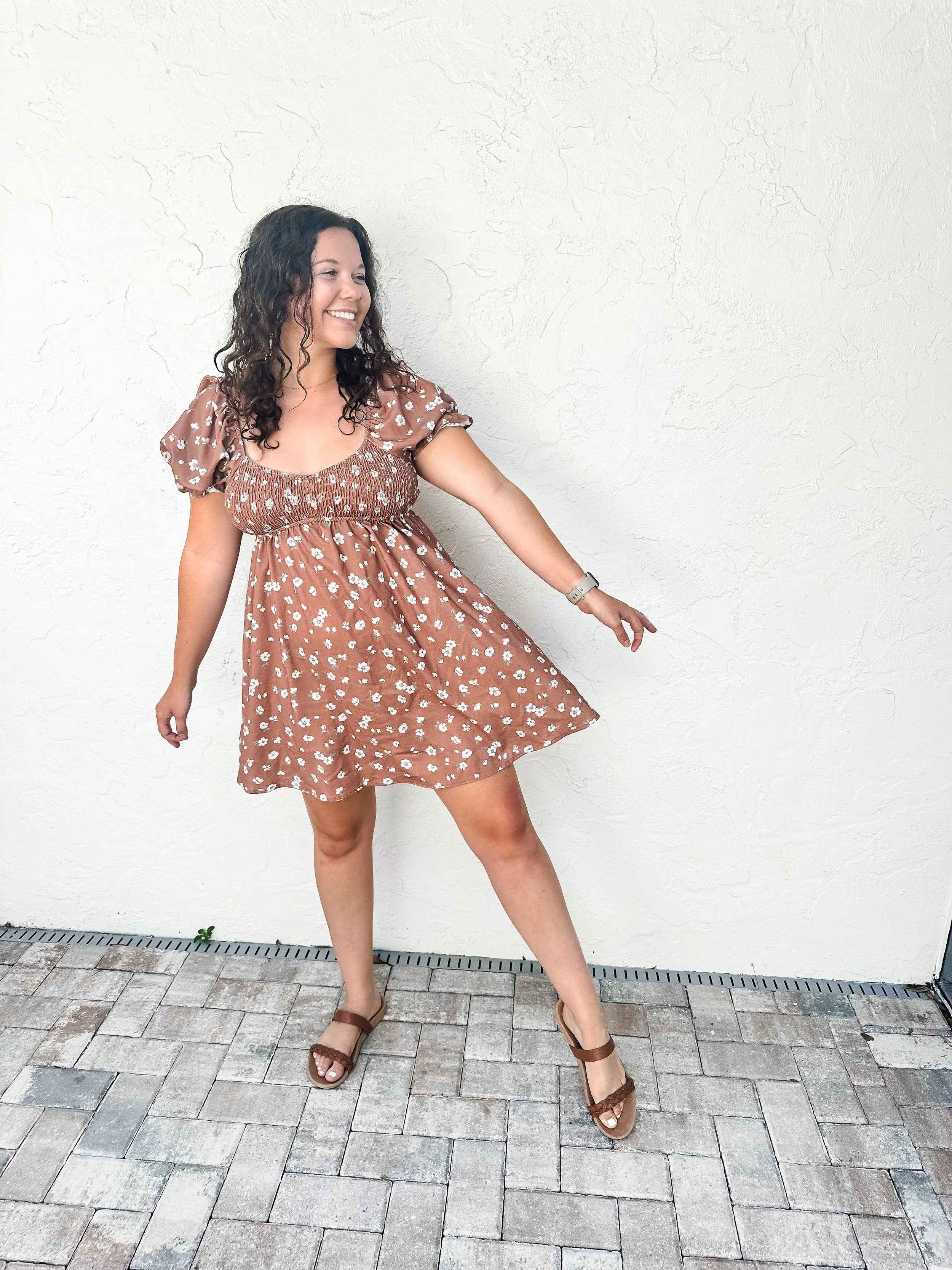 A woman in a brown floral dress with puffed sleeves and a cinched waist, smiling and posing against a white textured wall, with her right arm extended downward and her left arm bent, wearing brown sandals and an Apple Watch.