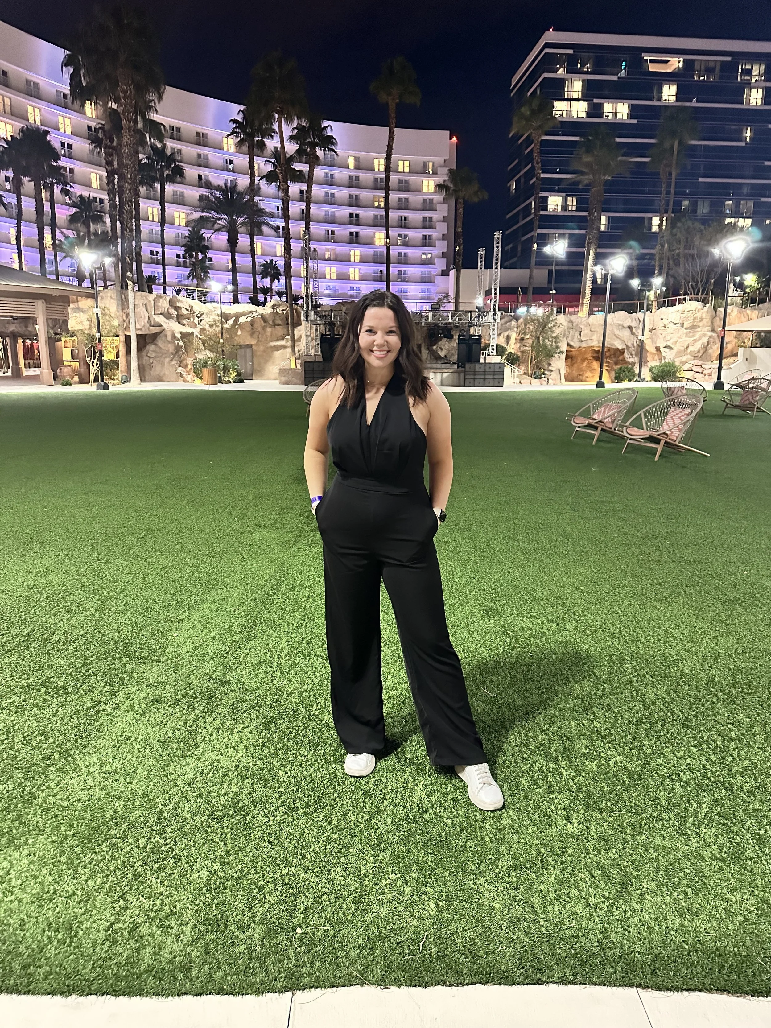 A woman in a black sleeveless jumpsuit and white sneakers standing on a well-lit artificial grass area at night with modern buildings, palm trees, and outdoor seating in the background.