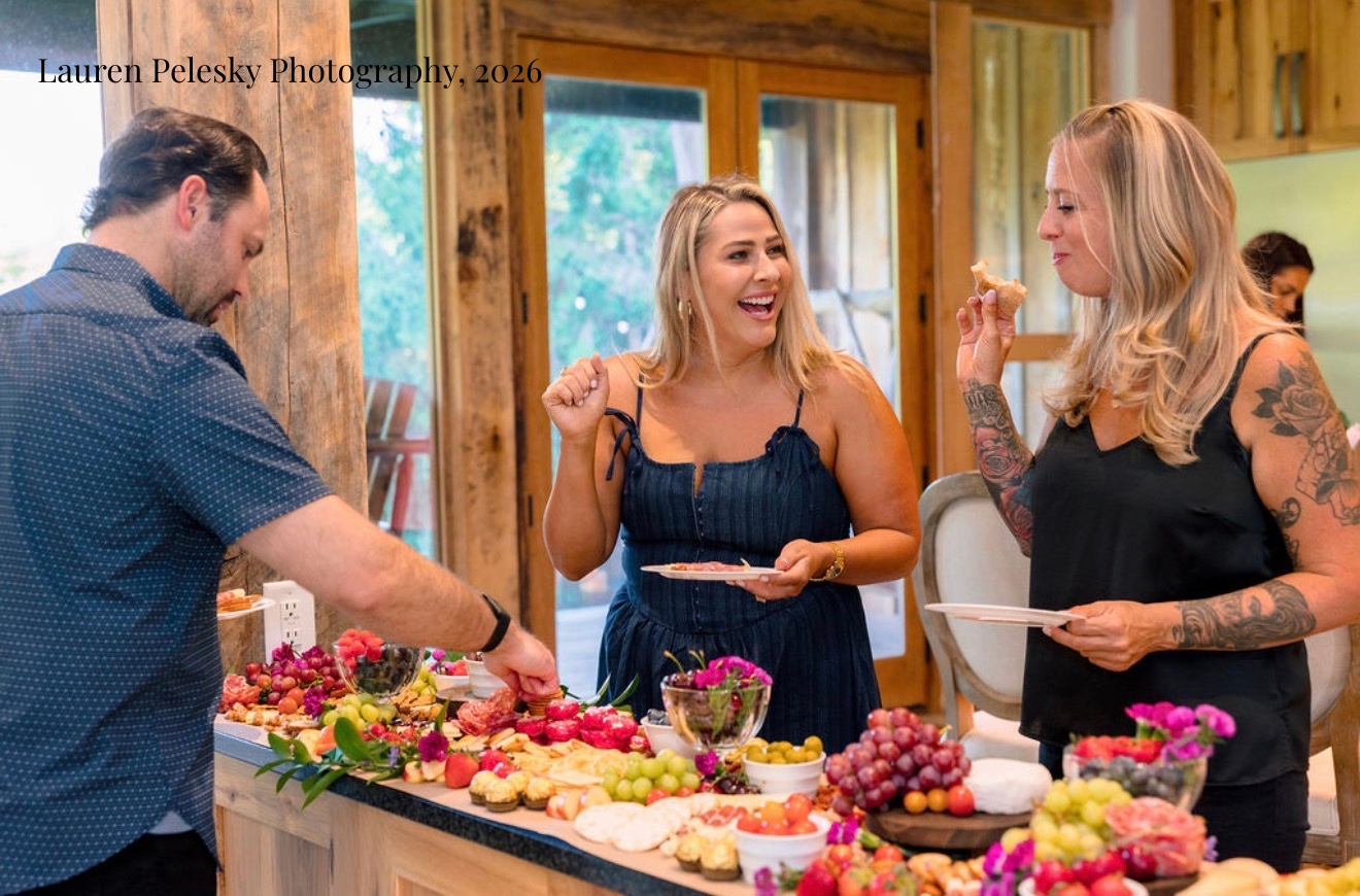 Wedding guests laugh together while enjoying an artistic grazing table filled with cheeses, fruits, meats, and other snacks.